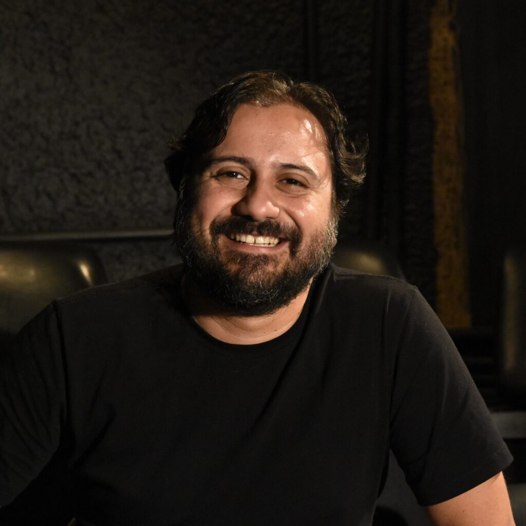 Homem branco, sorrindo, com barba e cabelos castanho escuros. está usando uma blusa preta.